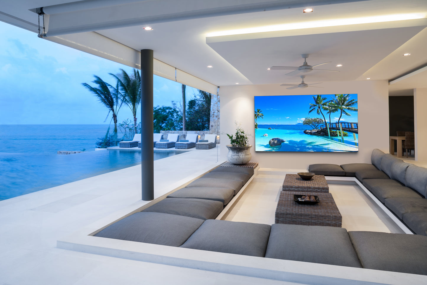 A modern outdoor living room with a low sectional sofa, a woven coffee table, and a large wall-mounted outdoor TV.